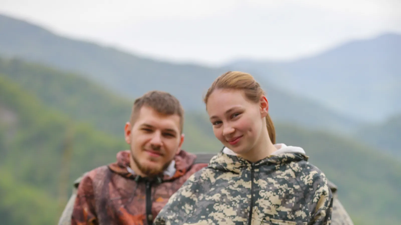 Пара в защитных куртках спокойно позирует на квадроцикле на фоне гор, шлем закреплён на руле